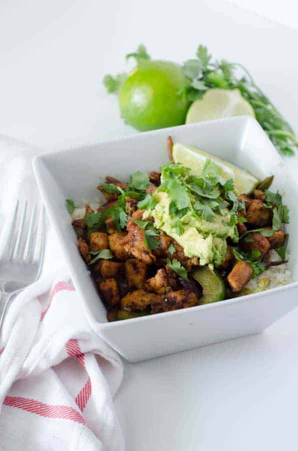 homemade chipotle burrito bowl with cauliflower rice, peppers, onions, and chicken topped with avocado and cilantro, with sliced lime and cliantro in the background