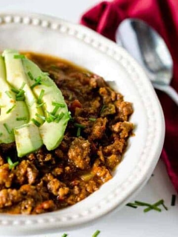 white bowl full of chili topped with avocado slices with red napkin and spoon next to it