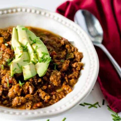 white bowl full of chili topped with avocado slices with red napkin and spoon next to it