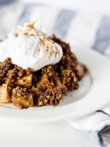 paleo apple cobbler on a white plate with a dollop of coconut whipped cream on top and a blue and white striped towel in the background