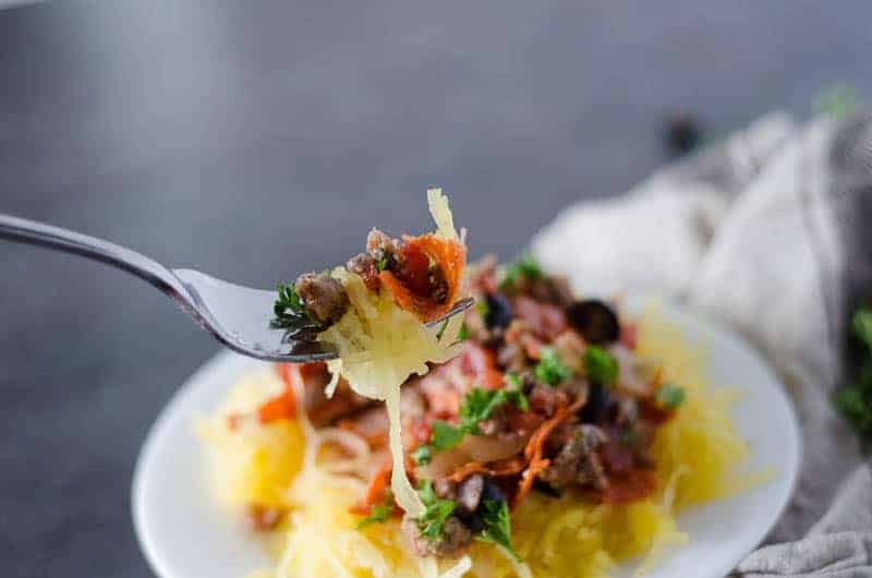 Fork containing a bite of paleo spaghetti squash pizza pasta with a blurred plate of food in the background