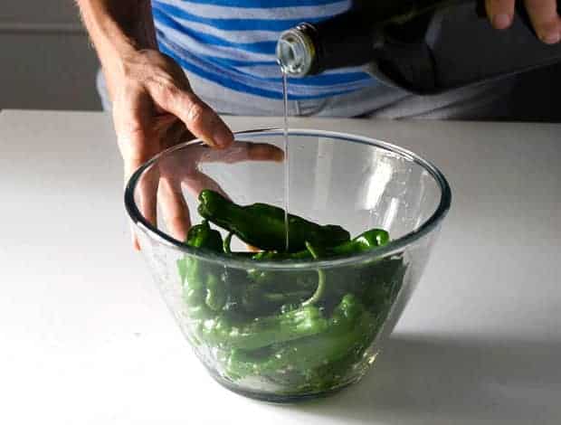 Side view of hand holding a bowl of shishito peppers pouring oil into the bowl