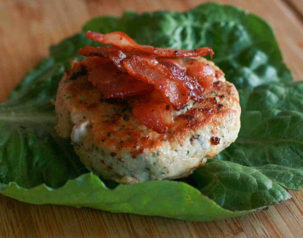 side view of a chicken burger topped with bacon on a lettuce leaf