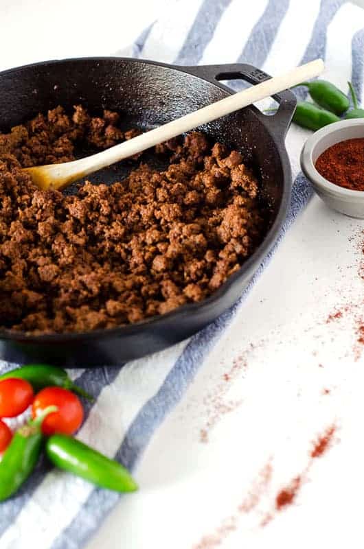 vertical image of cast iron skillet with taco meat and a wooden spoon on a blue and white striped dish towel