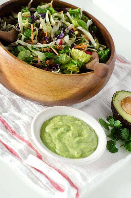 Whole30 taco salad on table with bowl of avocado dressing surrounded by avocado and cilantro