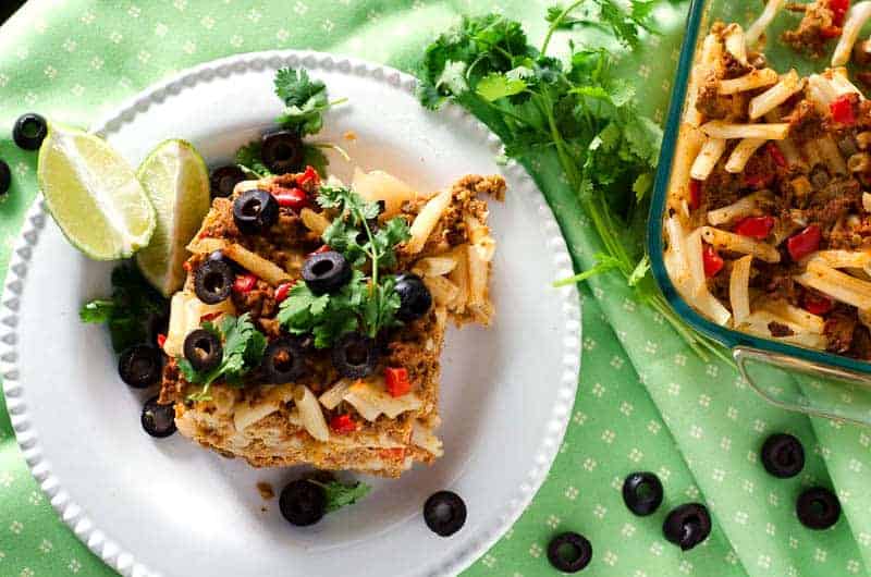 Overhead view of serving of chicken taco casserole on a white plate surrounded with cilantro, lime slices, and black olives