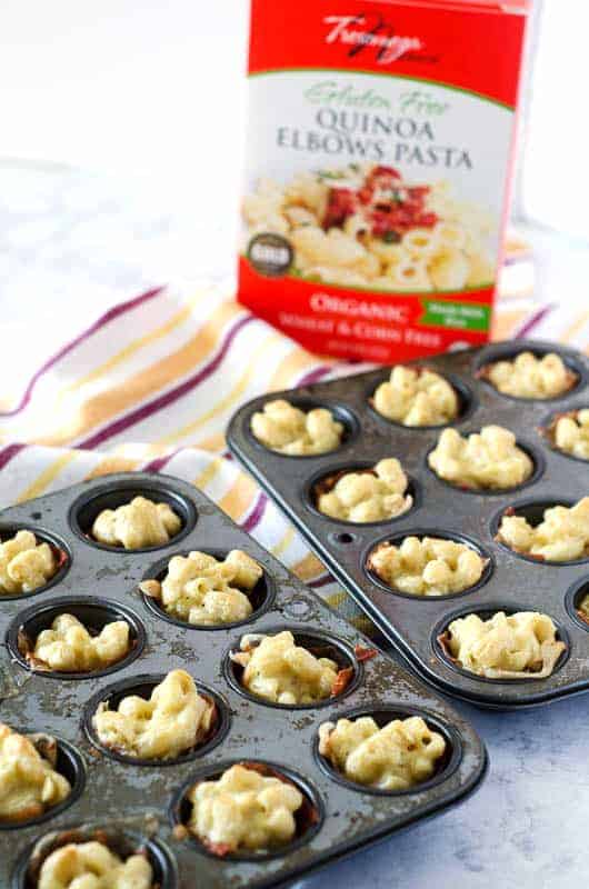 two mini muffin tins lined with proscuitto and filled with gluten free mac and cheese with box of quinoa pasta in the background