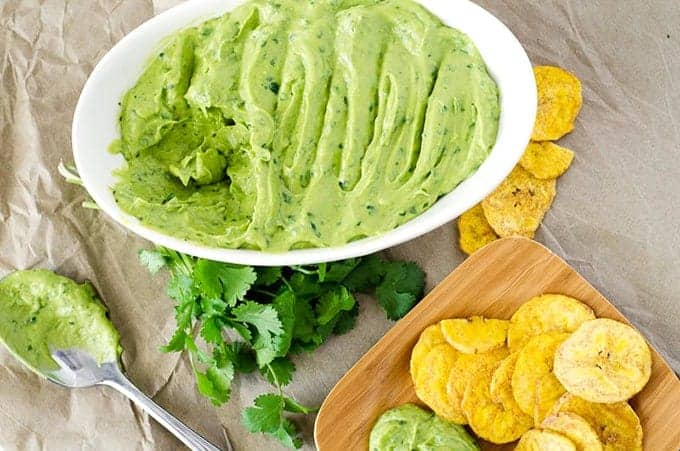 Overhead view of bowl of creamy avocado dip surrounded by spoon, loose cilantro, and plate of chips