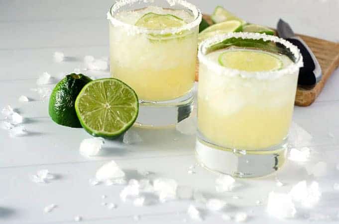 side angle view of two salt-rimmed paleo margaritas in glasses surrounded by limes and ice