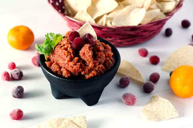 side view of black bowl full of cranberry orange salsa with a basket of chips in the background