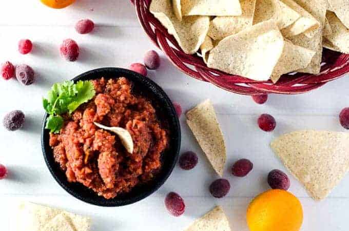 overhead view of a bowl of cranberry orange salsa and a basket of chips surrounded by cranberries and chips