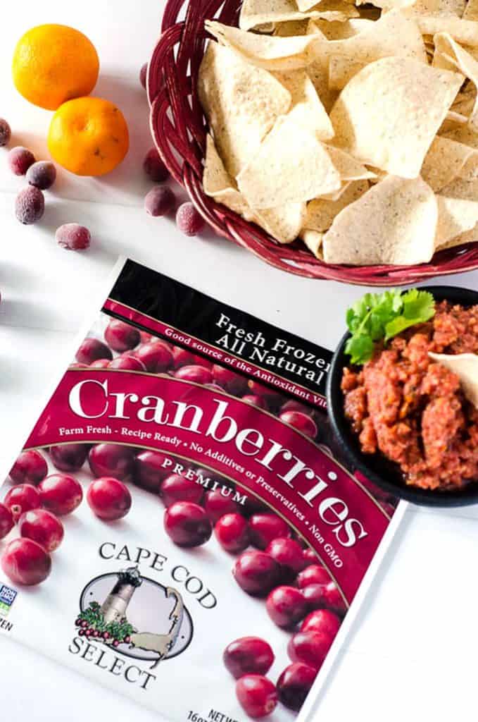 overhead view of a bowl of cranberry orange salsa, a basket of chips, and frozen cranberry package