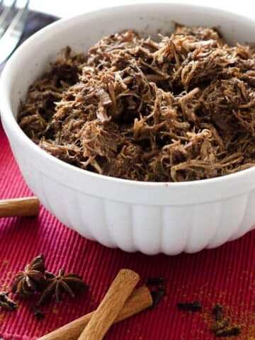 white bowl of pulled pork surrounded by whole spices on a red placemat