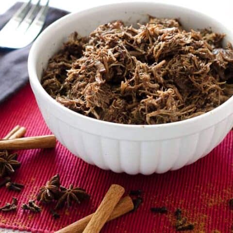 white bowl of pulled pork surrounded by whole spices on a red placemat