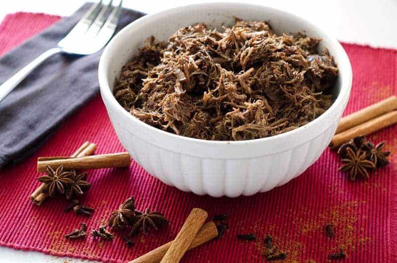 white bowl of pulled pork surrounded by whole spices on a red placemat