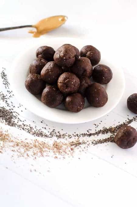 Plate of chocolate no-bake vegan energy balls surrounded by chia seeds, flax meal, and spoon with nut butter