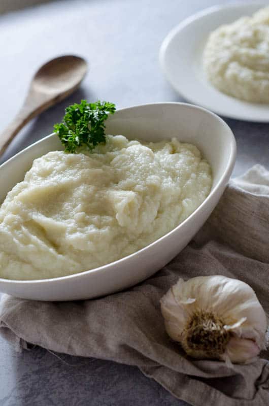 white oval bowl full of creamy cauliflower mashed potatoes on a napkin with a head of garlic and serving spoon