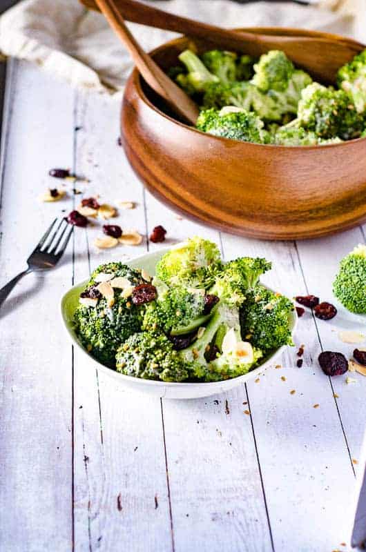 side view of white bowl with broccoli salad in it, with a wooden bowl full of the salad in the background and  fork on the table