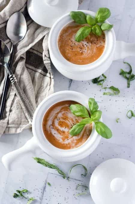 overhead view of two white crocks filled with tomato basil soup with spoons, lids and a napkin surrounding