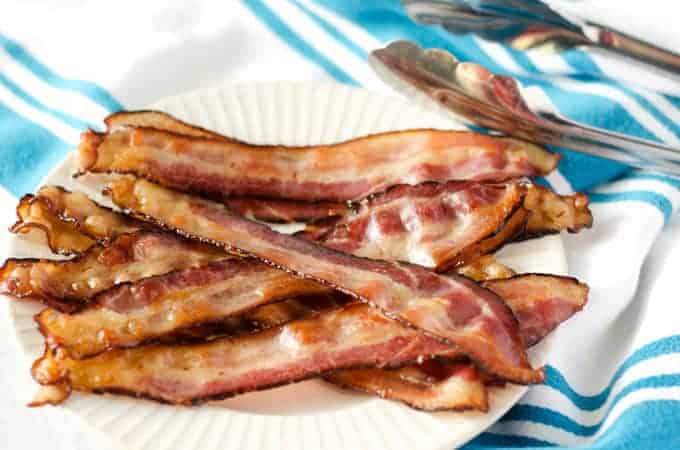 pile of bacon on a white plate on a white and blue striped towel with tongs in the background