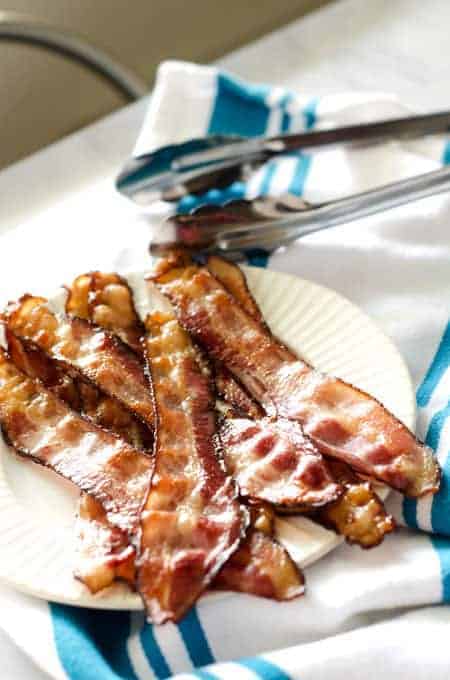 homemade bacon slices on a plate with tongs in the background