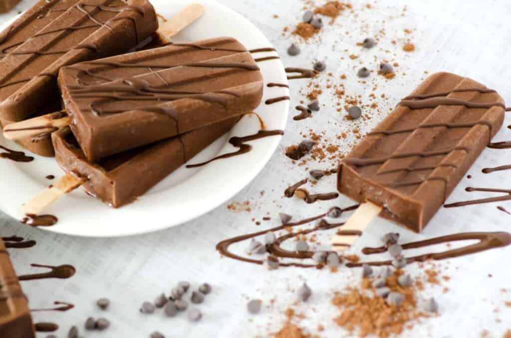 Side angle view of chocolate keto popsicles on a tabletop surrounded by cocoa powder, chocolate chips, and a chocolate sauce drizzle