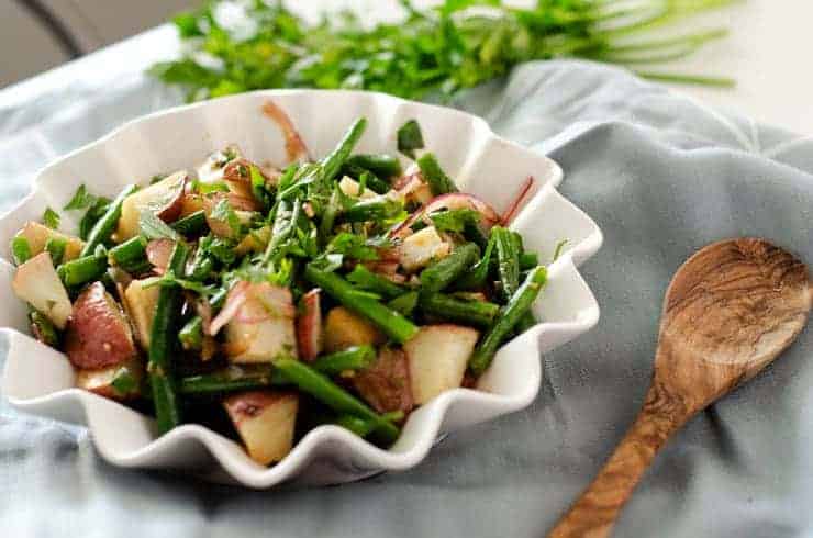 Green bean potato salad topped with fresh herbs in a white bowl with a wooden spoon next to it and fresh herbs in the background