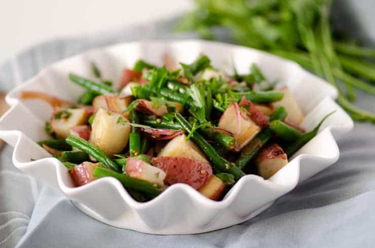 Close up of green bean potato salad in a white fluted edge bowl on top of a blue napkin with fresh herbs in the background