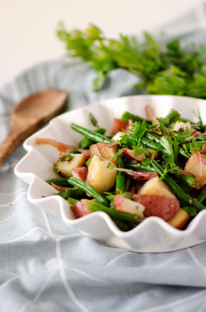 Green beans and red potatoes in a white fluted edge bowl on top of a blue napkin with wooden spoon in the background