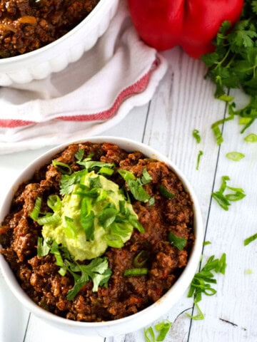 overhead view of a bowl of paleo chili surrounded by chives and a red bell pepper in the background
