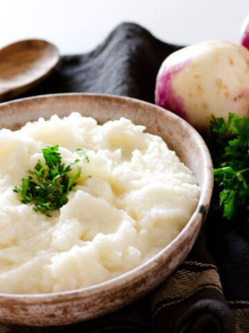 bowl of mashed turnips topped with fresh parsley with whole turnips in the background