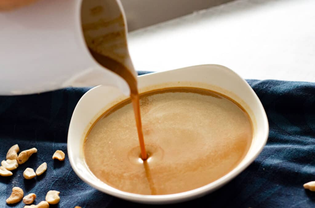 white bowl of pad thai peanut sauce on a dark blue napkin with white pitcher pouring sauce into bowl