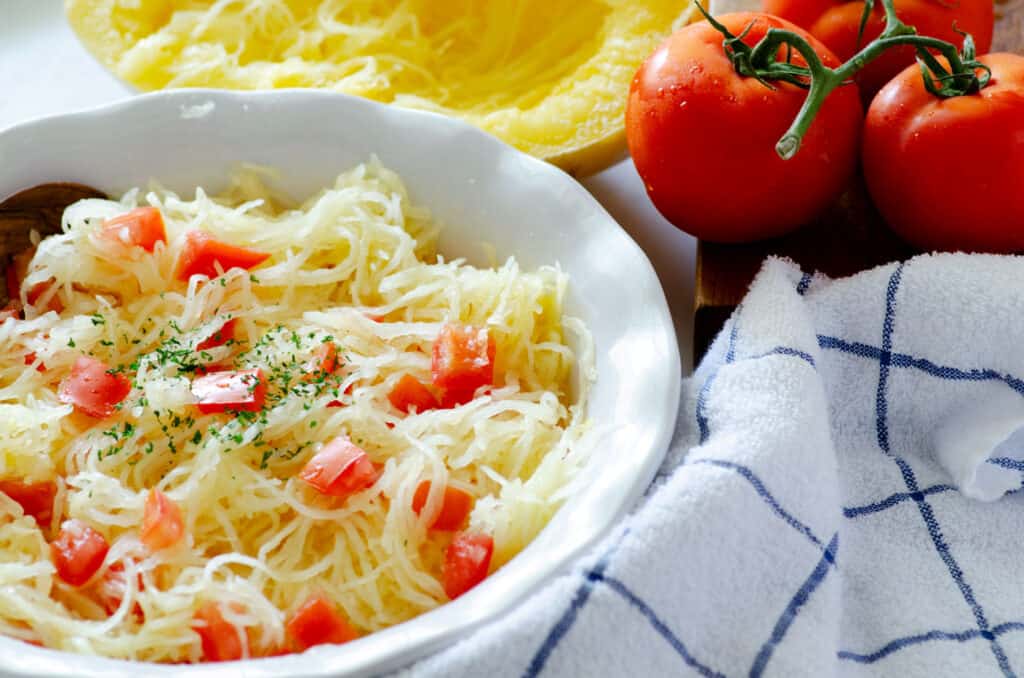 dish of spaghetti squash surrounded by a dish towel, tomatoes, and half a spaghetti squash