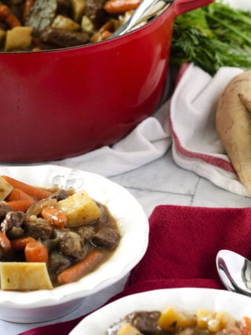 beef stew with carrots and potatoes in a white bowl with a red dutch oven in the background