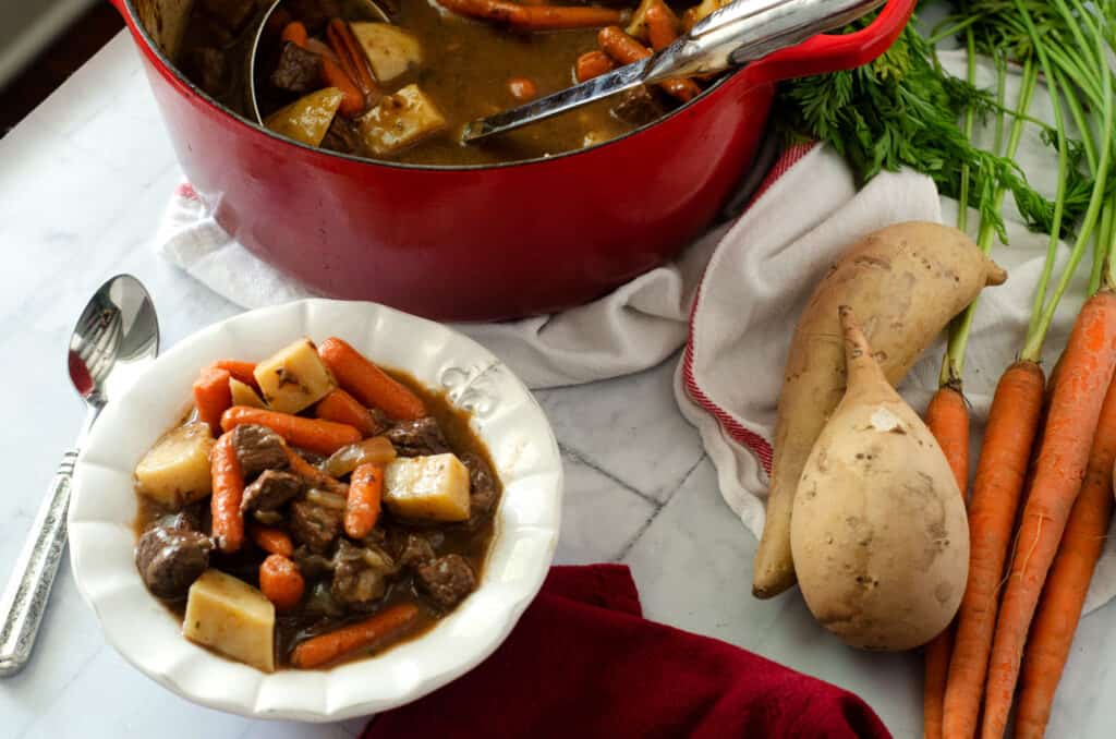 paleo beef stew in white bowl surrounded by sweet potatoes onions and dutch oven full of stew