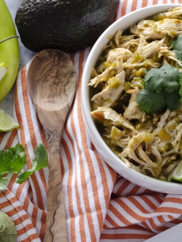 overhead view of bowl of green chile chicken surrounded by limes, avocadoes, and an orange striped towel