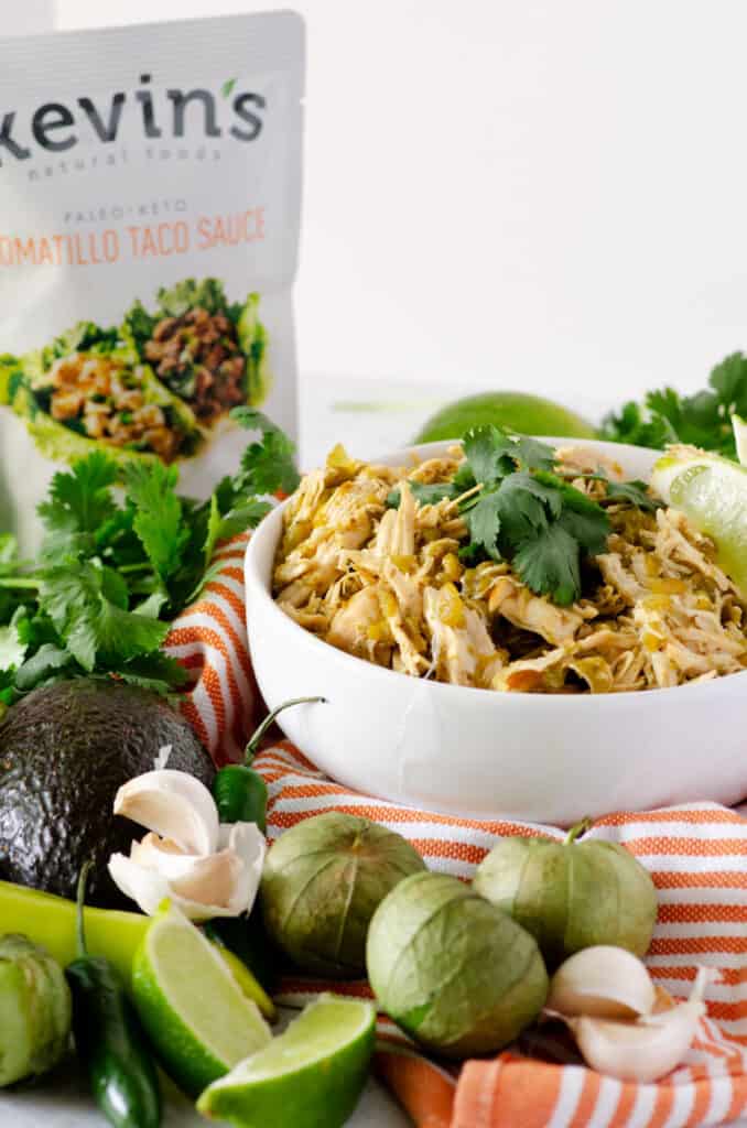 Bowl of green chile chicken surrounded by green produce and sitting on an orange and white striped towel