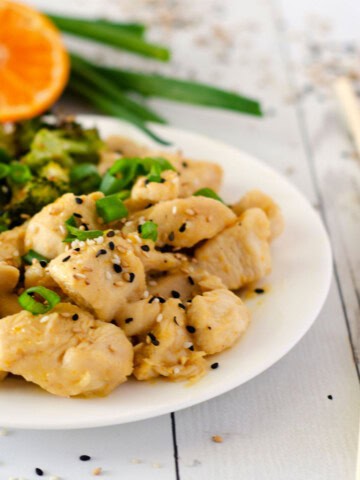 orange chicken on a white plate with vegetables in the background and chopsticks next to it