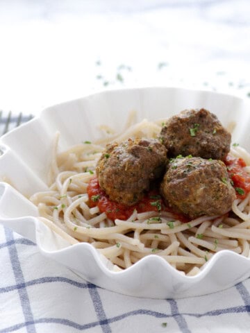 three paleo meatballs on top of red sauce and grain free spaghetti with a blue and white dishtowel