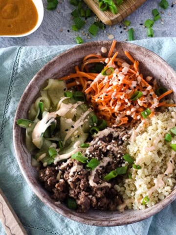 bowl filled with carrots, ground beef, cauliflower rice, and thin cucumber ribbons on top of a blue napkin with a wooden spoon next to it