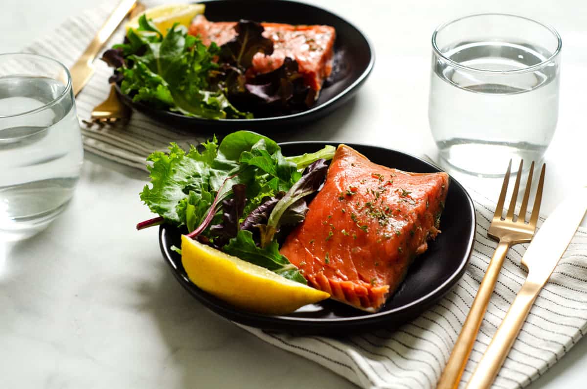 table setting of two plates of sous vide salmon with salad greens, lemon wedges and silverware