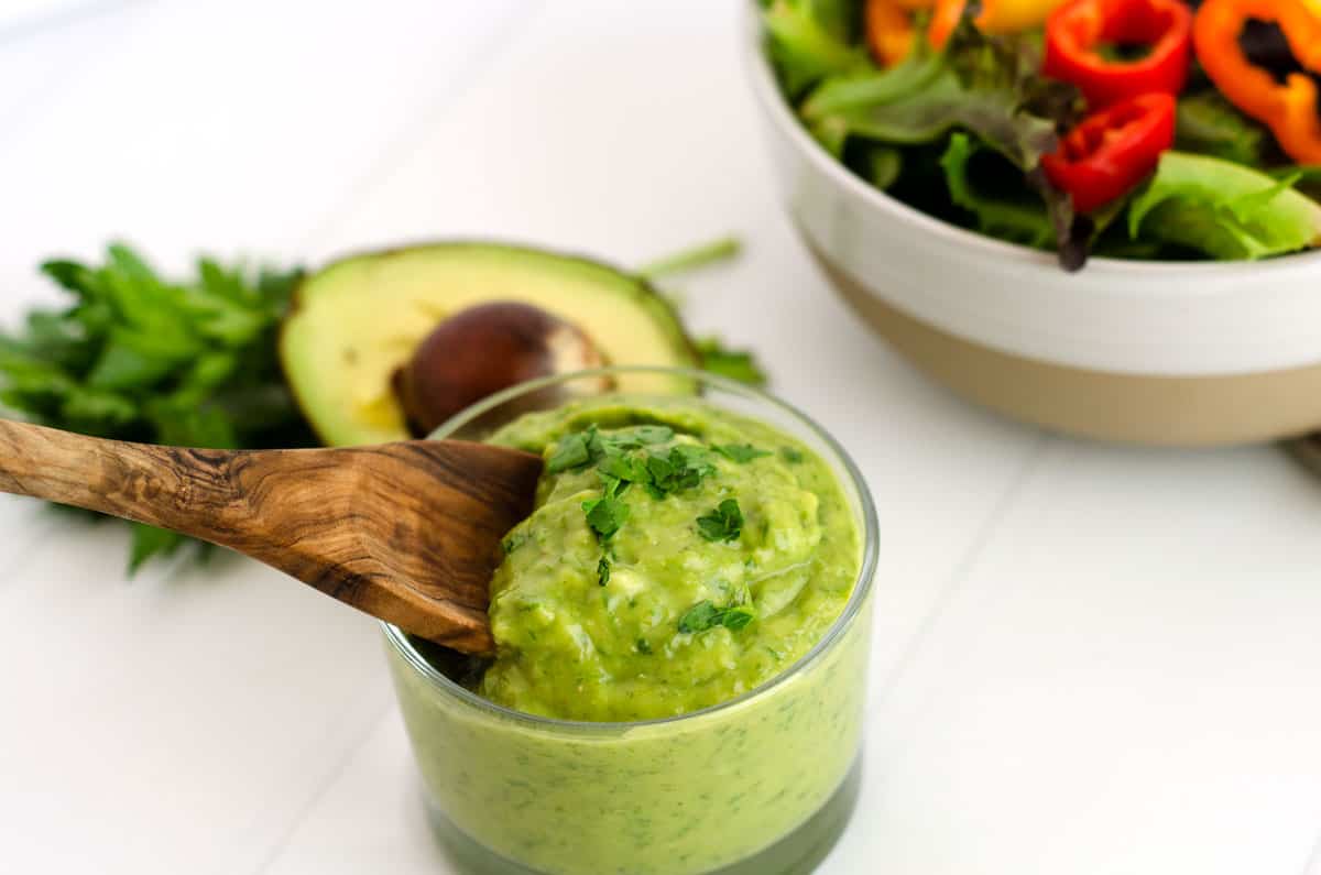 small glass bowl of homemade trader joe's green goddess dressing with a wooden spoon and a salad in the background