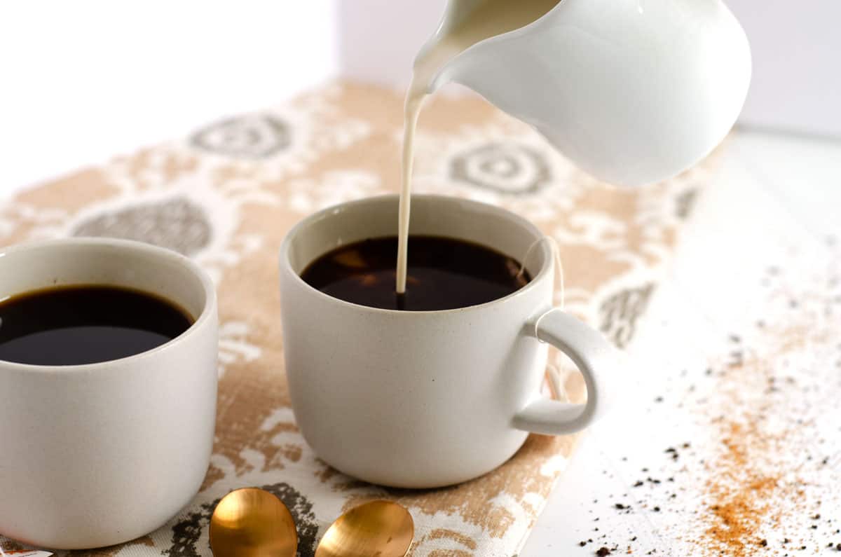 Side view of two dirty chai lattes with creamer being poured into one