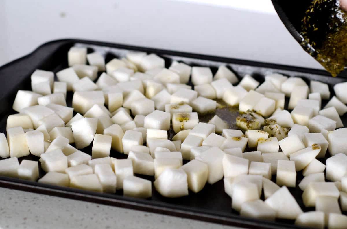 Baking sheet of raw turnip cubes with oil and spices being poured on top