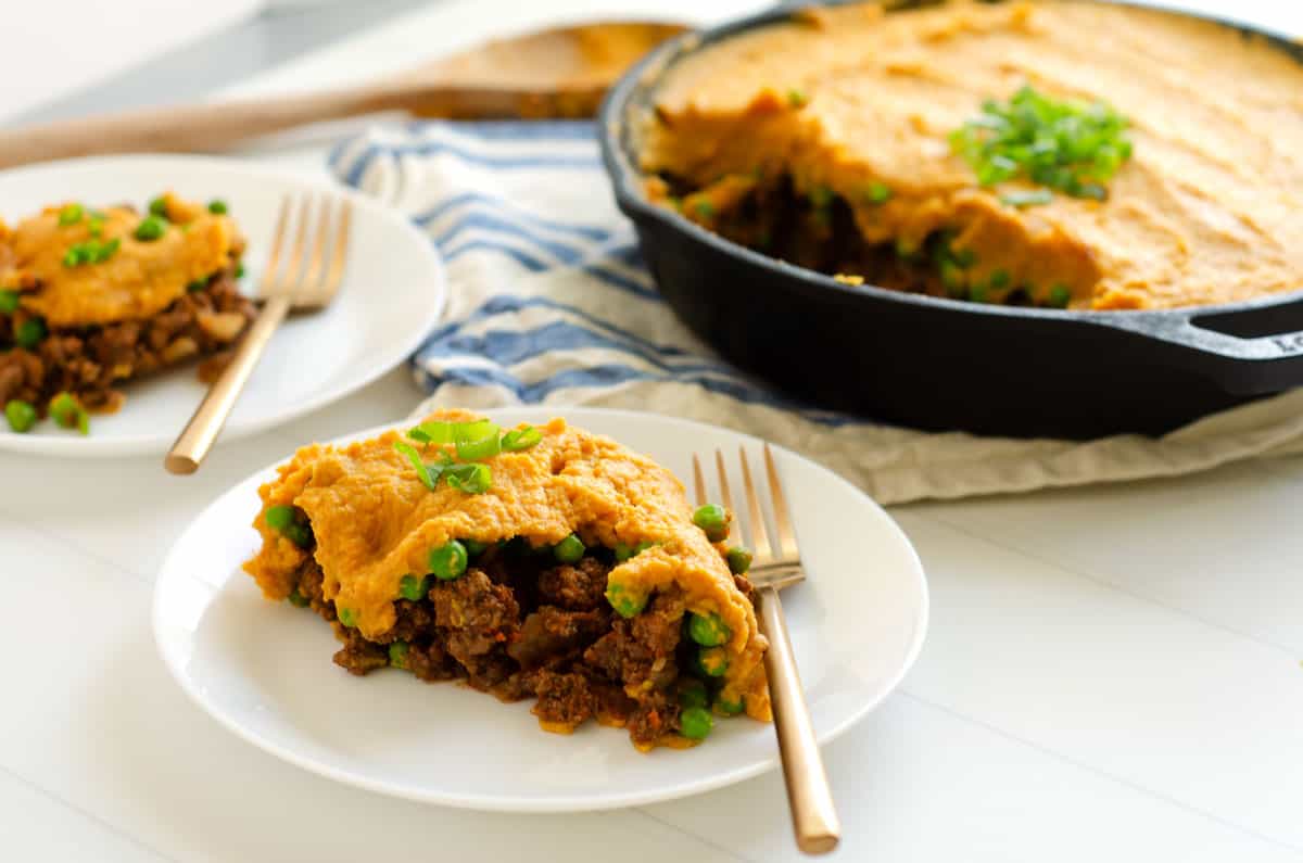 Two servings of shepherd's pie on white plates with gold forks with cast iron skillet in background