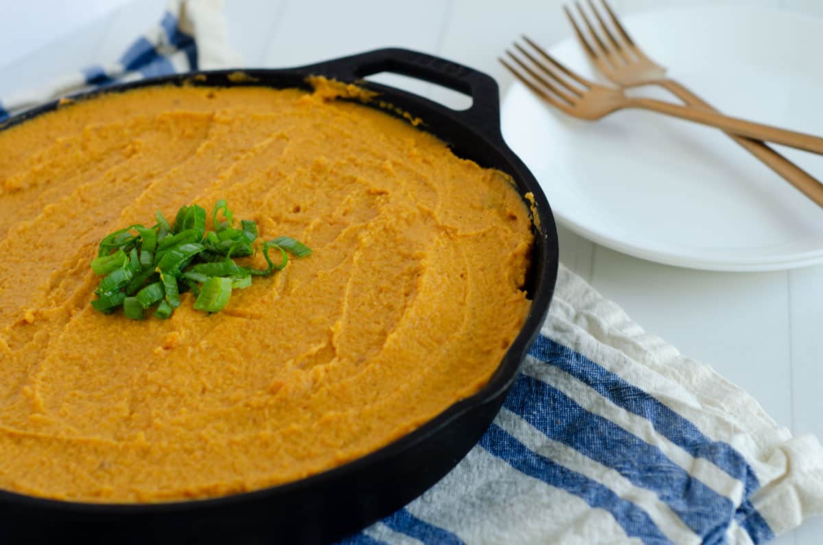 Cast iron skillet of shepherd's pie with sweet potato on a blue striped dish towel with white plates and gold forks in the background