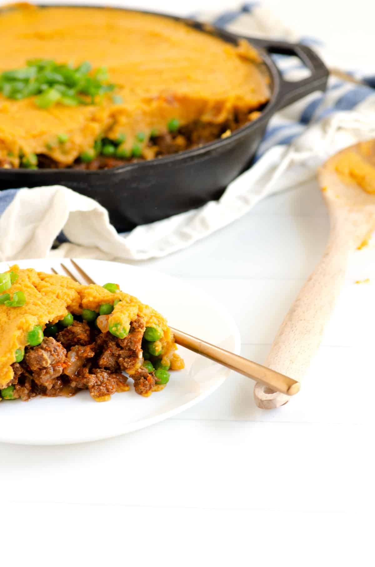 Serving of shepherd's pie with sweet potato on a white plate with a gold fork and cast iron skillet of shepherd's pie in background