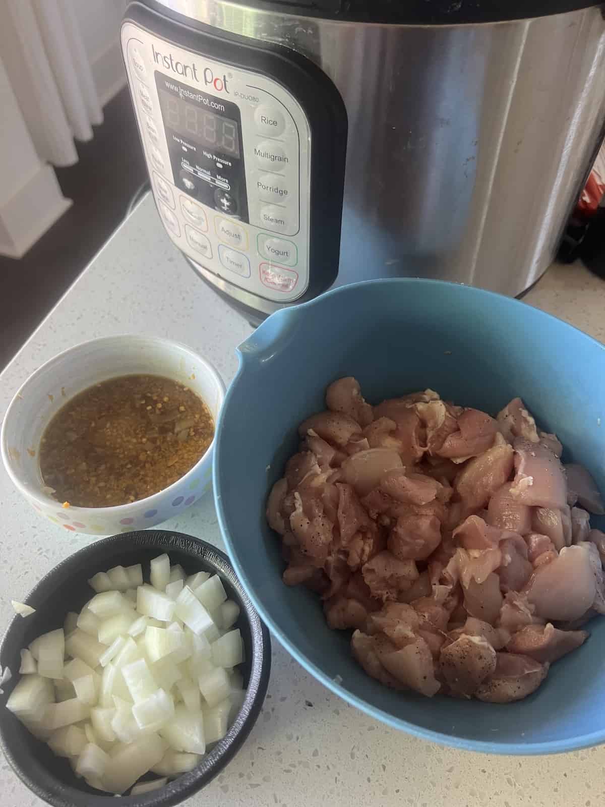 Instant pot on counter with bowl of raw chicken pieces, bowl of chopped onion, and bowl of orange sauce