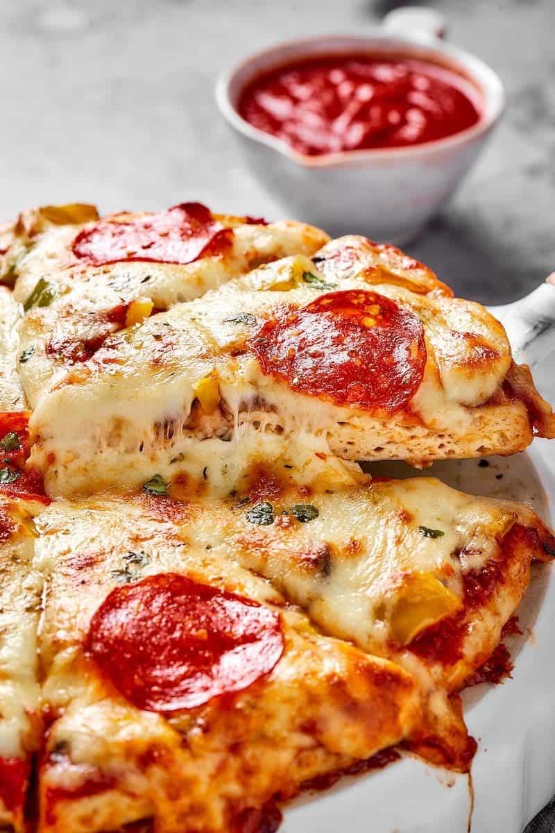 Chicken crust pizza slice being lifted off platter from pizza with cup of tomato sauce in the background