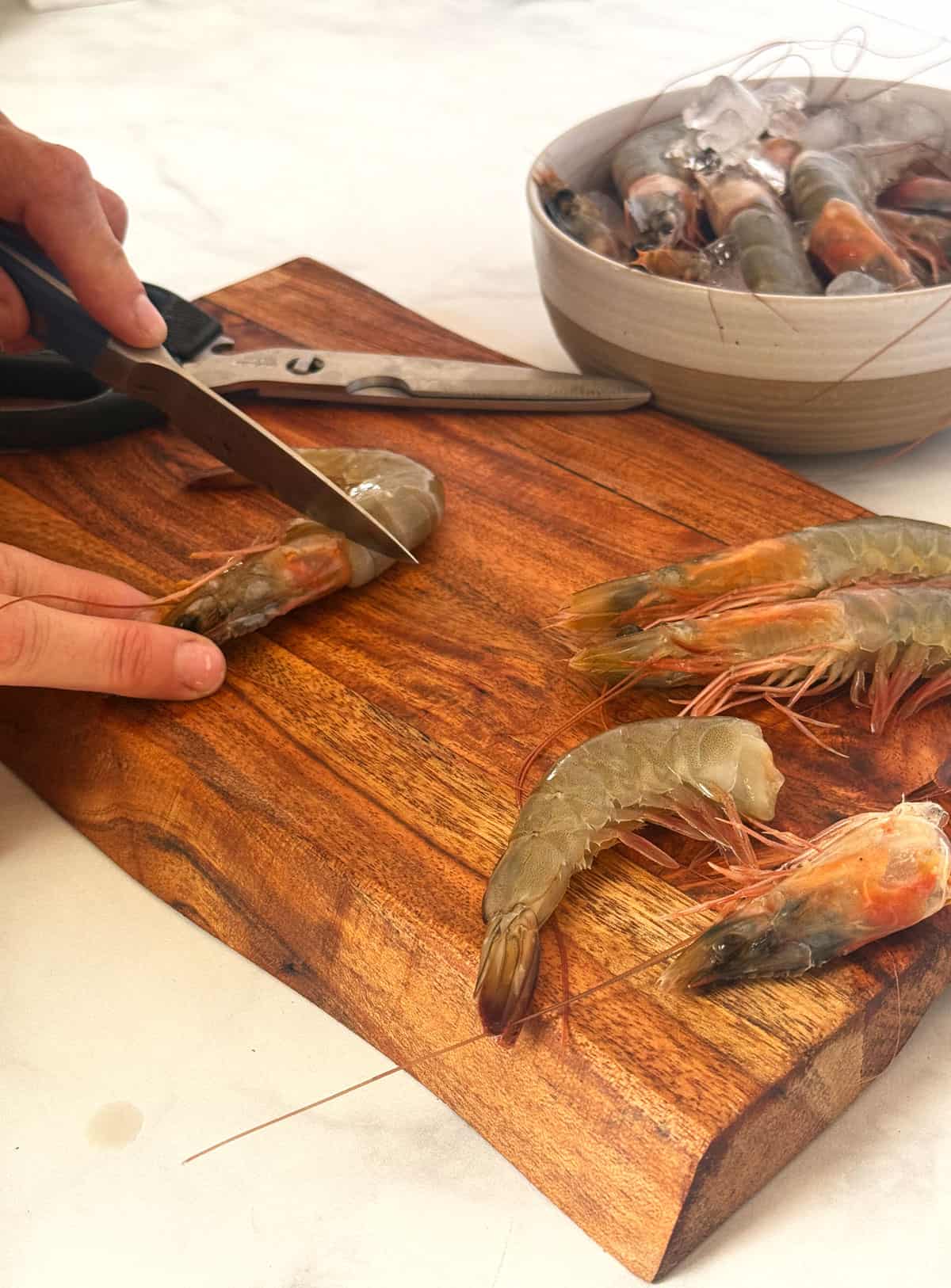 close up of a wooden cutting board with a bowl of head on shrimp in the background and a woman's hands removing a shrimp head with a paring knife
